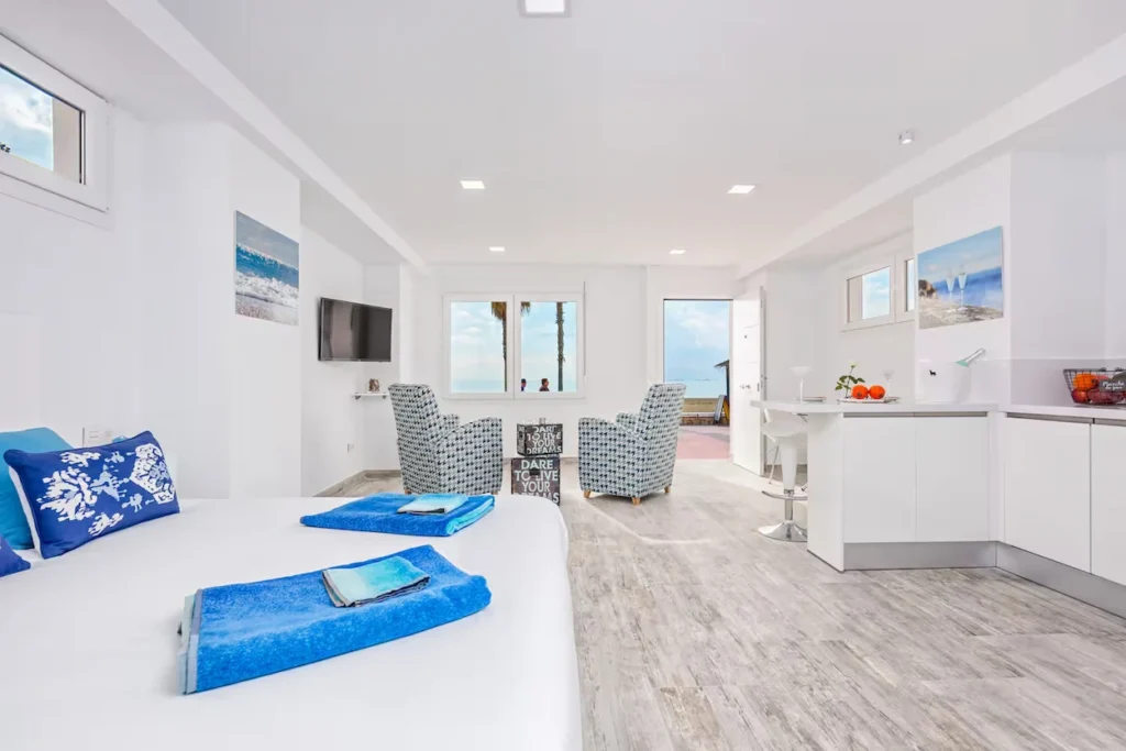 Bright modern studio apartment with white walls, blue accents, patterned chairs, and beach views