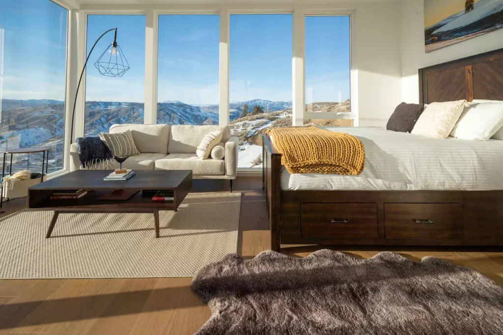 Bright bedroom with a dark wood bed frame, a small sofa, and floor-to-ceiling windows showing mountains