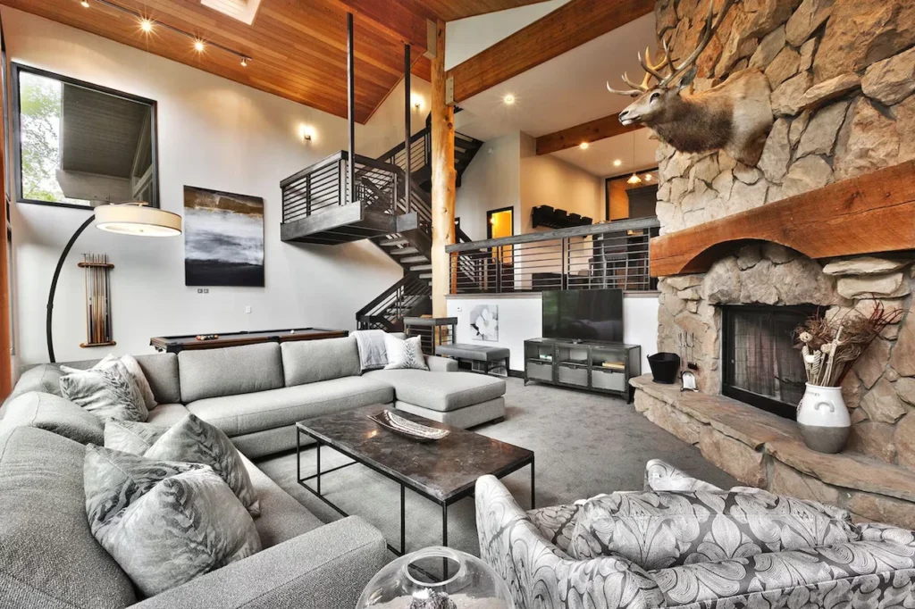 A spacious cabin living room with a grey sectional sofa, stone fireplace, mounted elk head, and pool table