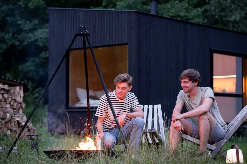 Two young men sitting in wooden chairs outside a black modern tiny cabin in the forest, tending to a campfire at dusk