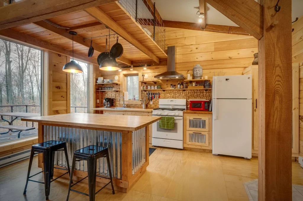 Cabin kitchen with a corrugated metal island, black barstools, a white refrigerator, and large windows
