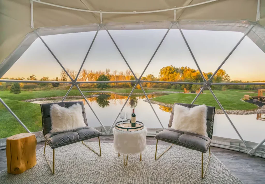Inside a glamping dome looking out at a pond through a geometric window with two gray accent chairs