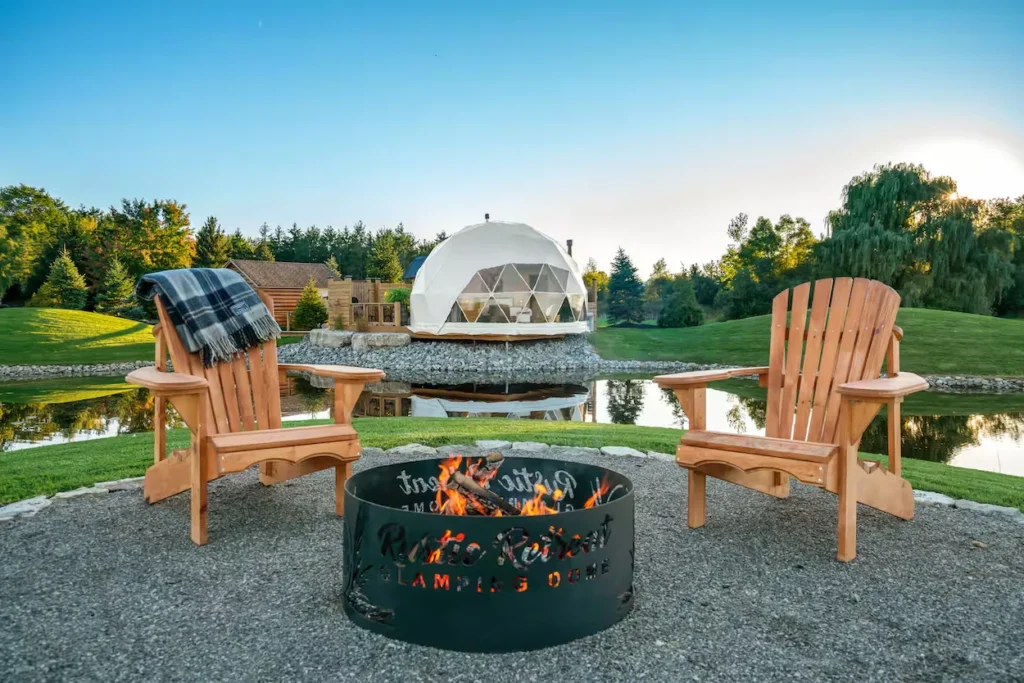 Two wooden Adirondack chairs by a burning fire pit overlooking a pond and white glamping dome