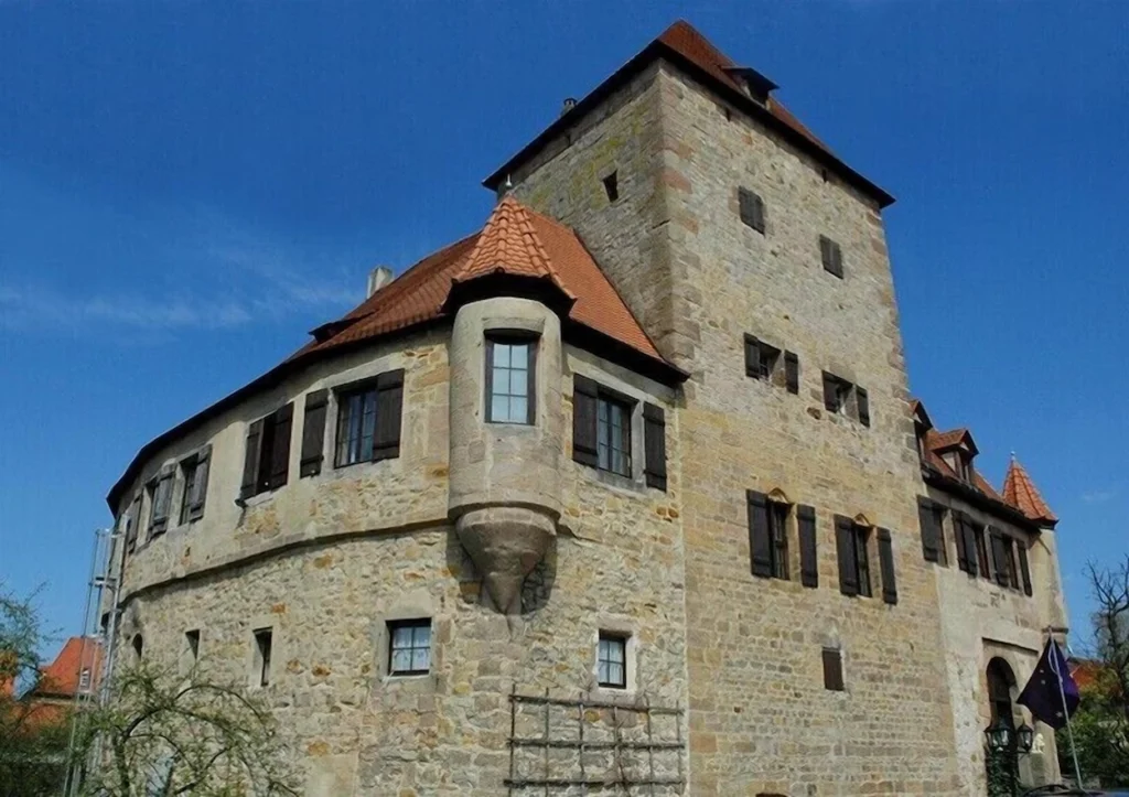 Historic stone castle with a large tower, shuttered windows, and a terracotta tiled roof under a blue sky