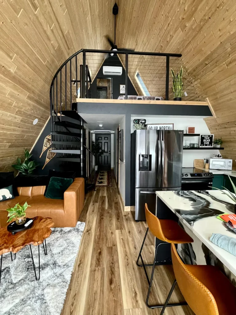 A-frame cabin interior featuring a spiral staircase, loft bedroom, leather sofa, and stainless steel refrigerator