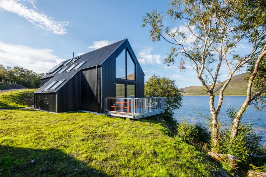 Modern black corrugated metal cabin with a deck on a grassy hill overlooking a lake