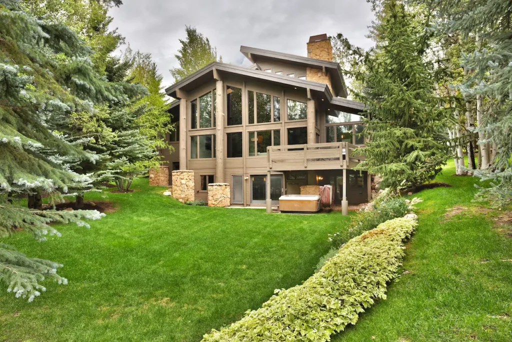 A modern multi-level mountain lodge with a large green lawn, hot tub, and surrounding evergreen trees