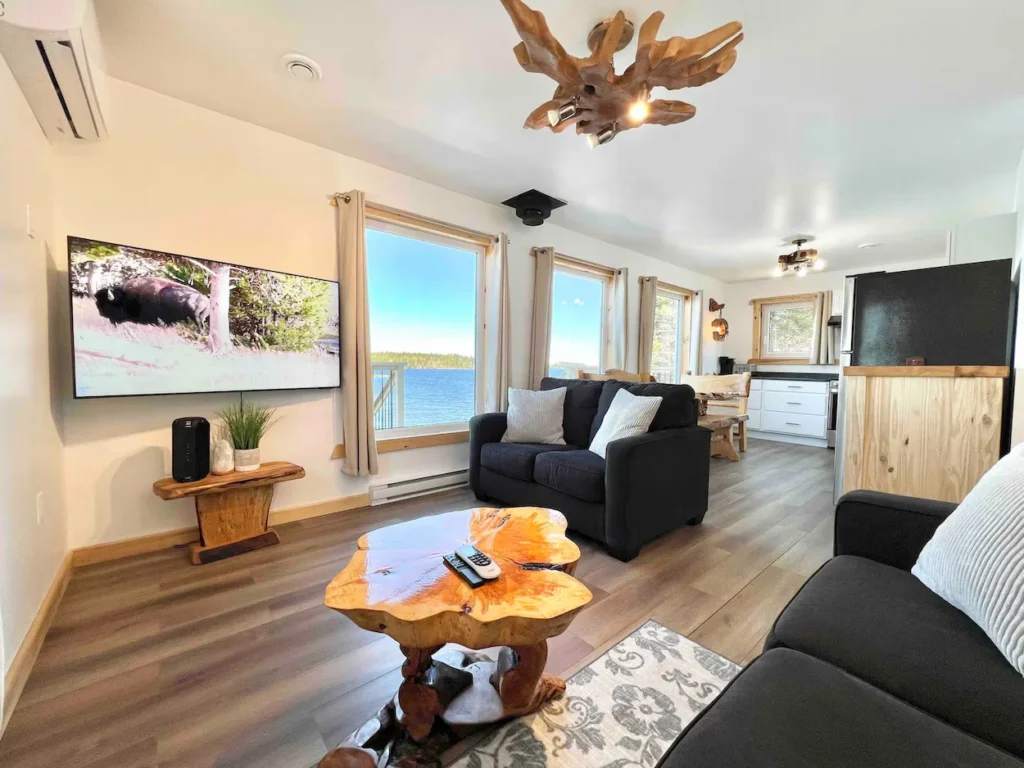 Cottage living room featuring dark sofas, a live edge wood coffee table, and large windows overlooking the lake