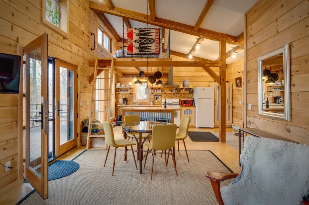 Log cabin interior with a round glass dining table, kitchen, and a ladder leading to a sleeping loft