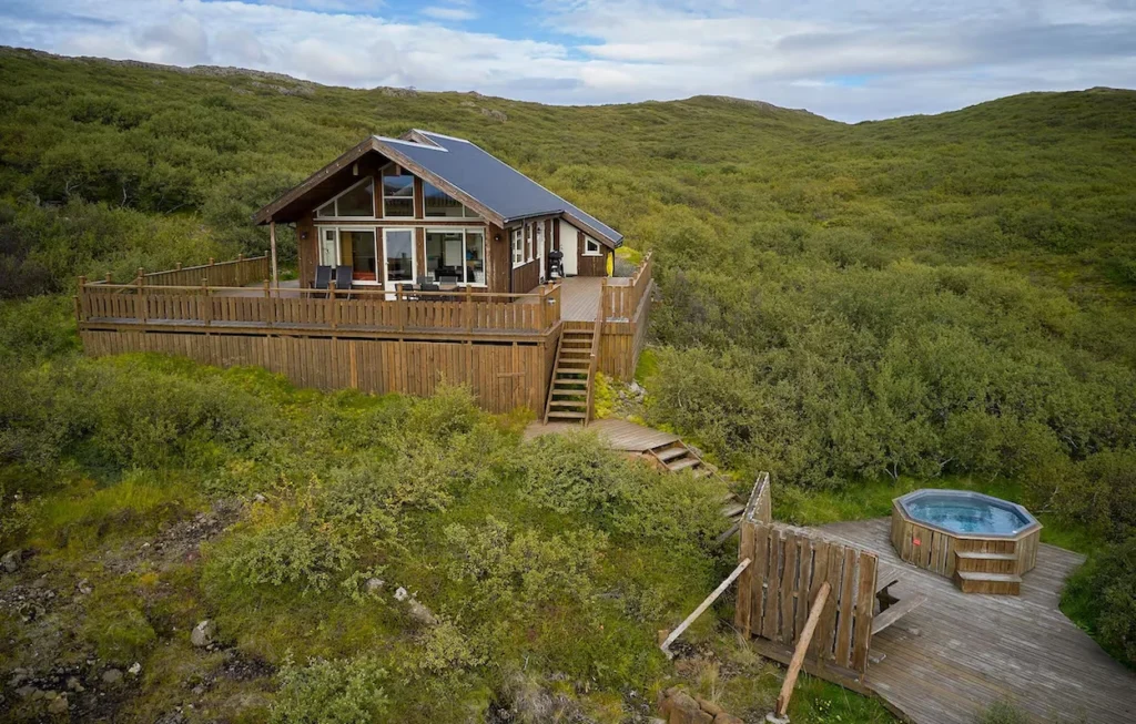 A wooden cabin on a grassy hill with a wrap-around deck and a separate hot tub