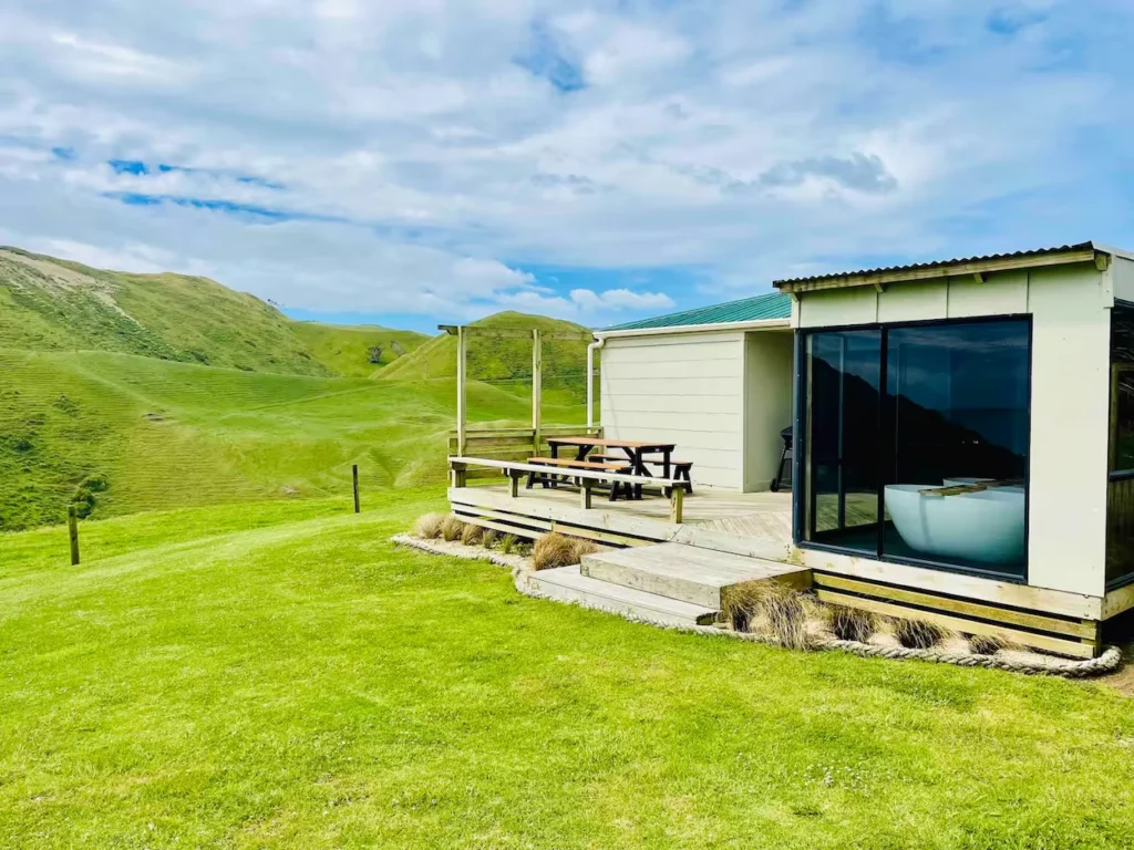 Modern white cottage with a glass-enclosed bathtub room and wooden deck on a green hillside in New Zealand
