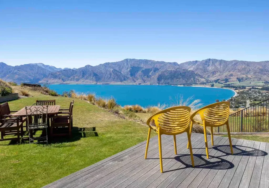 Yellow patio chairs on a wooden deck overlooking a bright blue lake and rugged mountains in New Zealand