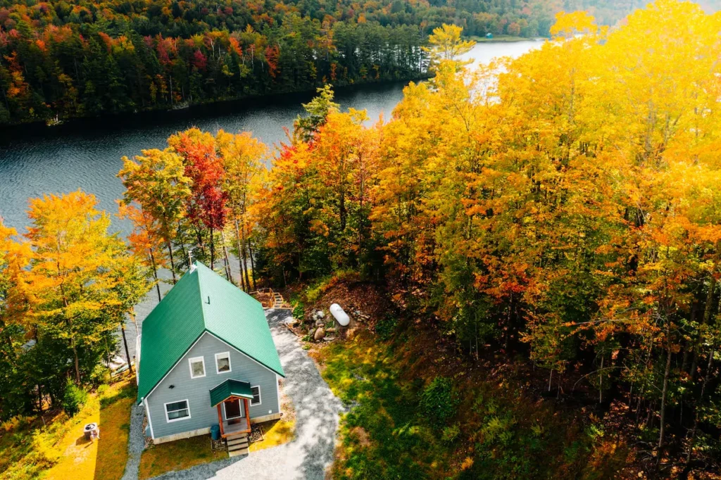 Aerial view of a gray lakefront cabin with a green tin roof surrounded by vibrant fall foliage