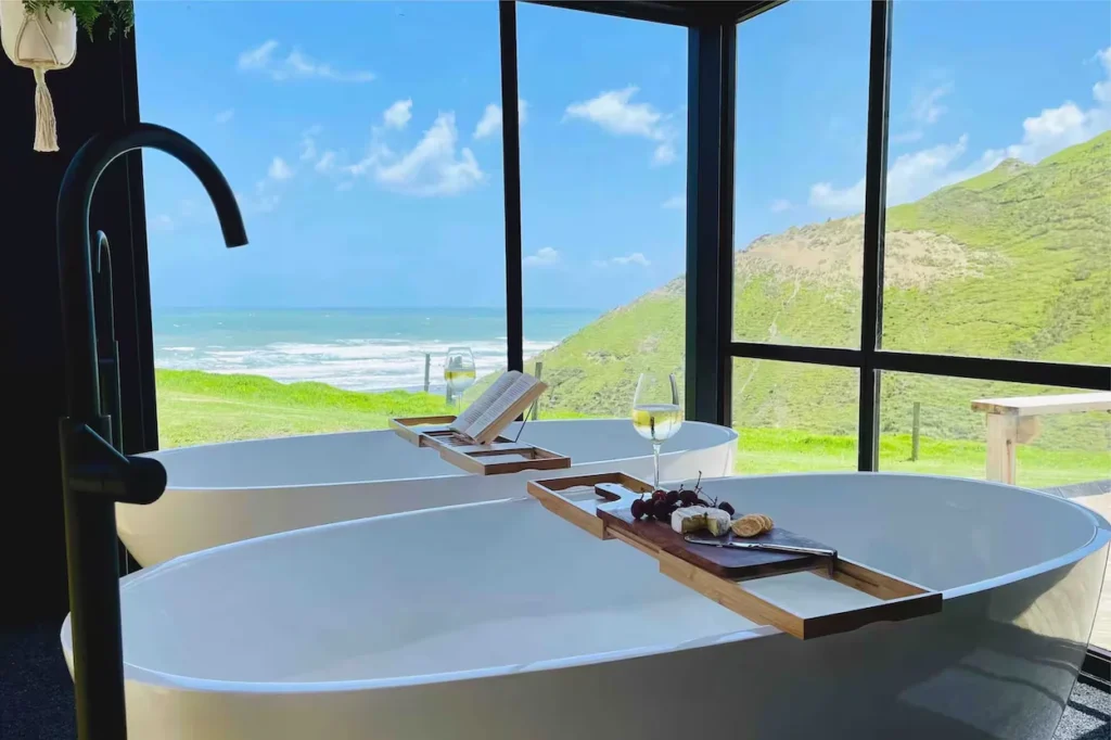 Two modern white bathtubs with wooden trays looking out floor-to-ceiling windows at the ocean and green hills