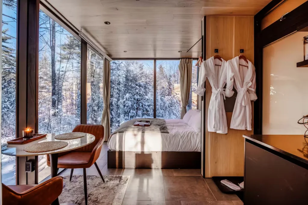 Glass cabin bedroom with a dining table and large window wall looking out at snow covered trees
