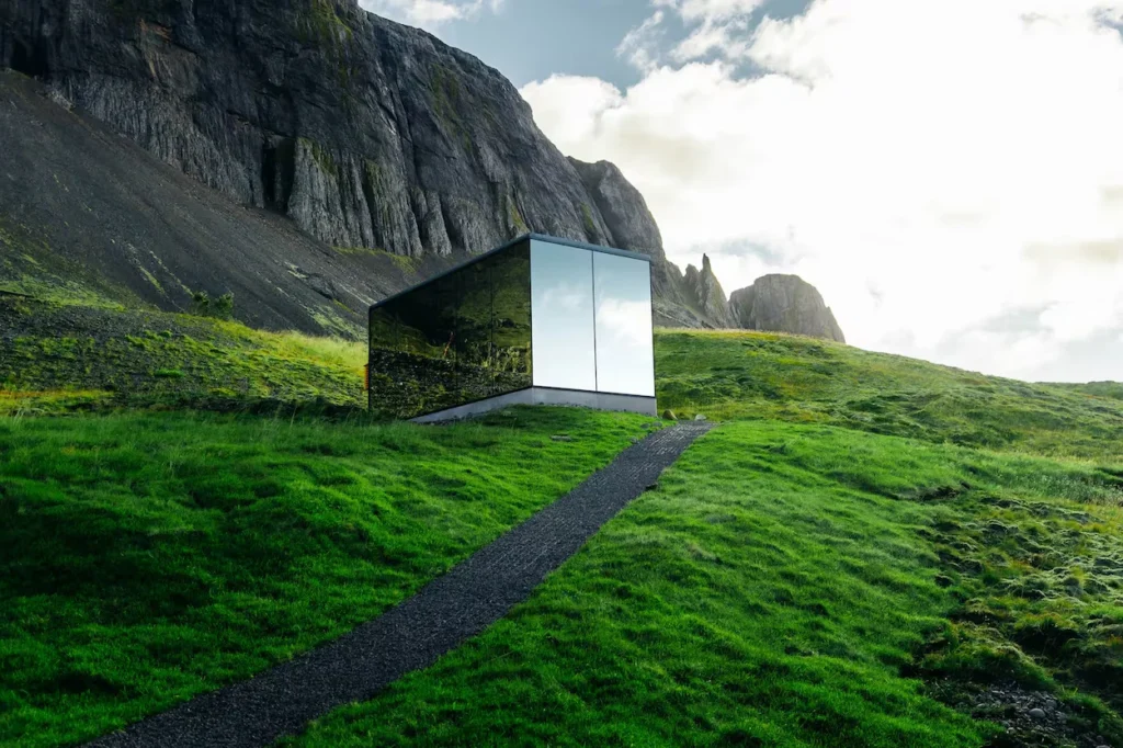 A modern cube-shaped mirror cabin sitting on a green hill beneath a rocky mountain cliff