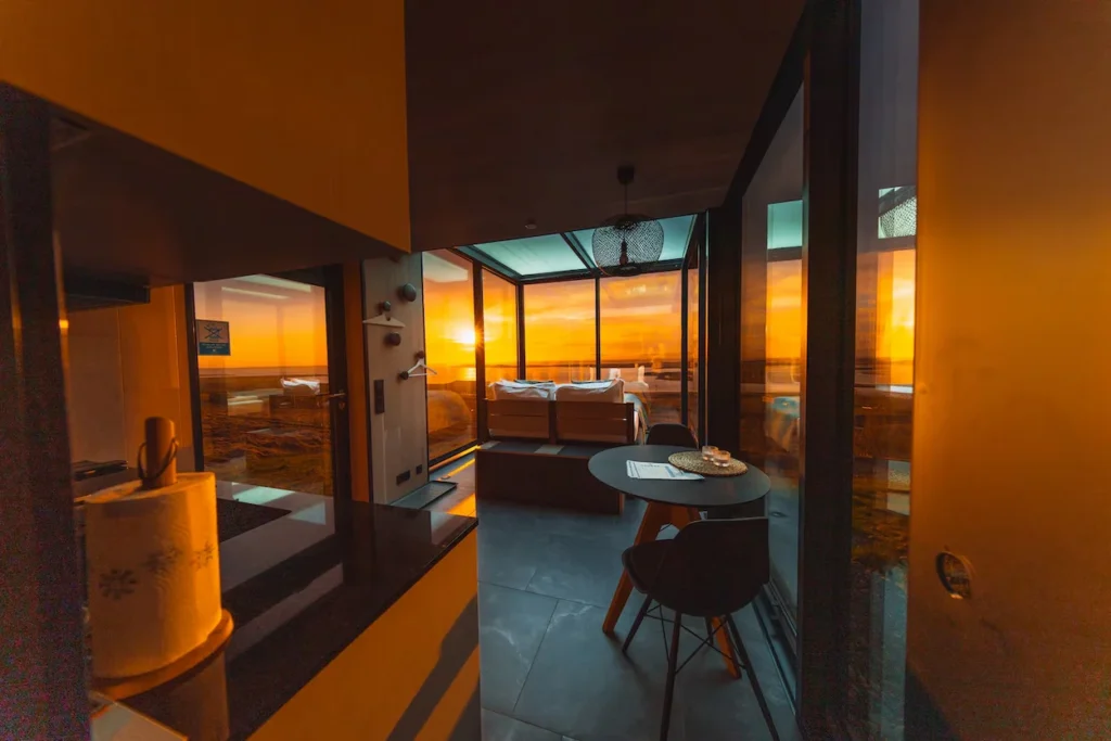 A modern glass cabin interior showing a dining table and bed against a bright orange sunset sky