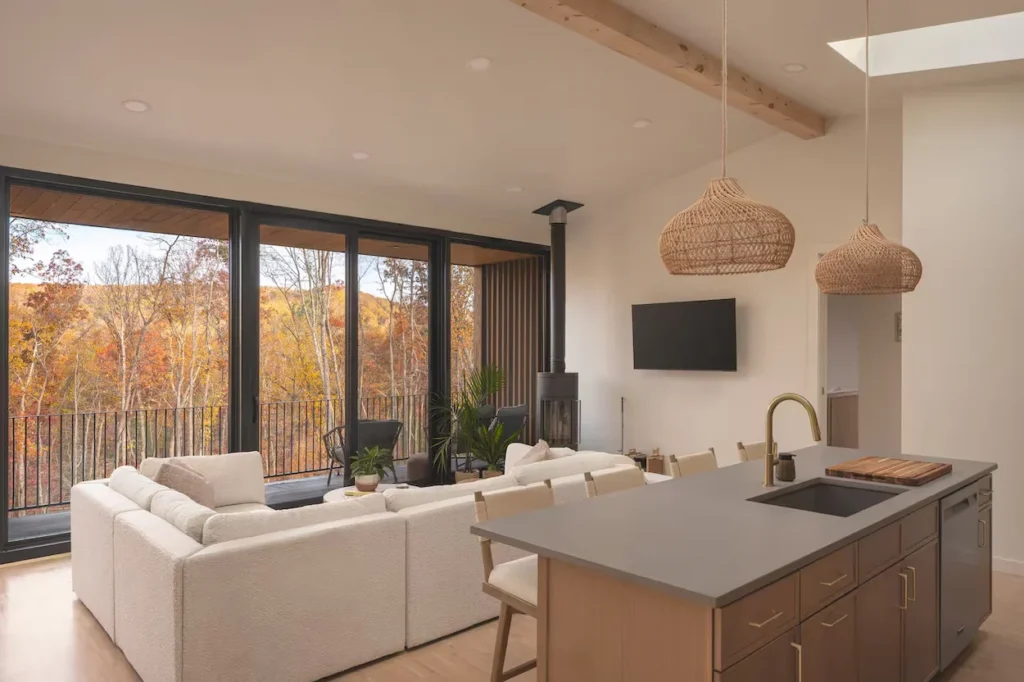 Minimalist cabin interior with a white sectional, kitchen island, and glass wall showing autumn trees