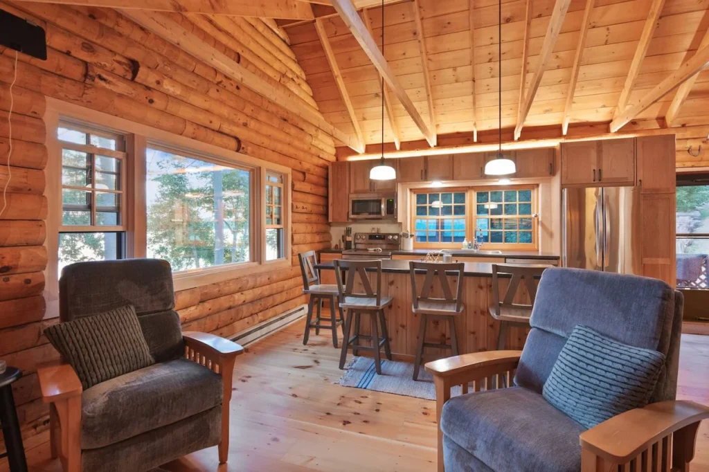 Log cabin interior showing a kitchen island with barstools next to a seating area with two armchairs