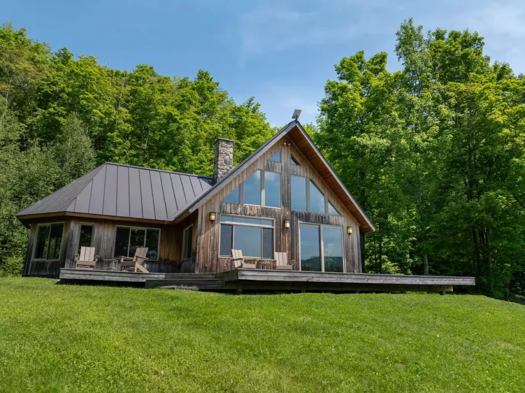 Wooden cabin with a stone chimney and large windows on a green lawn surrounded by trees