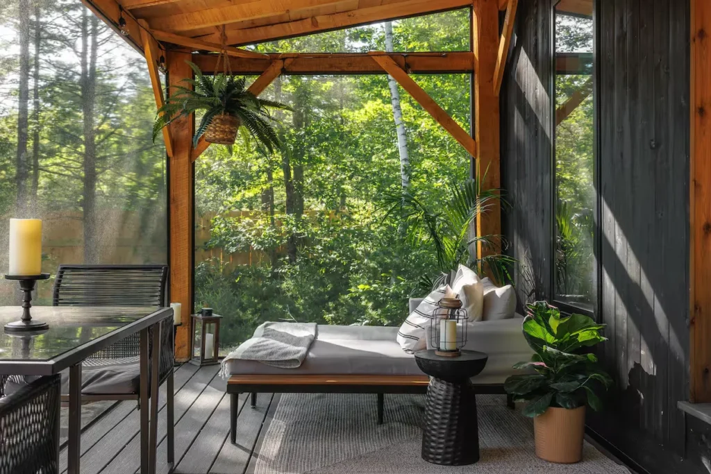 Screened porch with a white daybed, glass dining table, and potted plants overlooking green woods