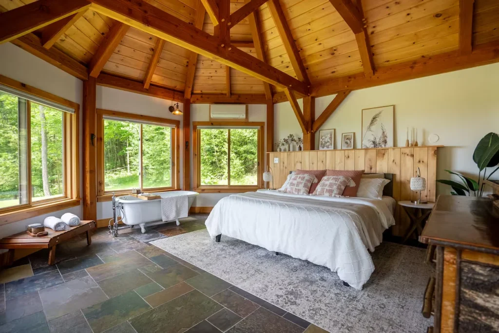 Cabin bedroom with vaulted wood ceilings, a slate tile floor, a bed, and a freestanding tub next to windows