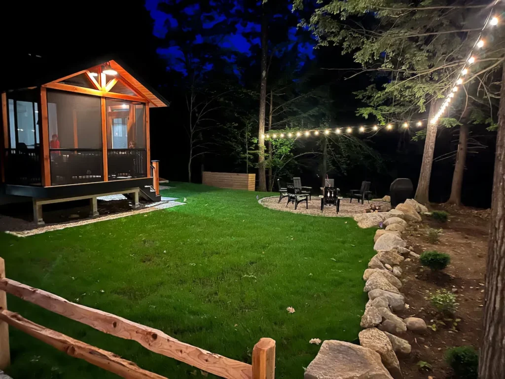 Cabin backyard at night with a green lawn, fire pit seating area, and glowing string lights