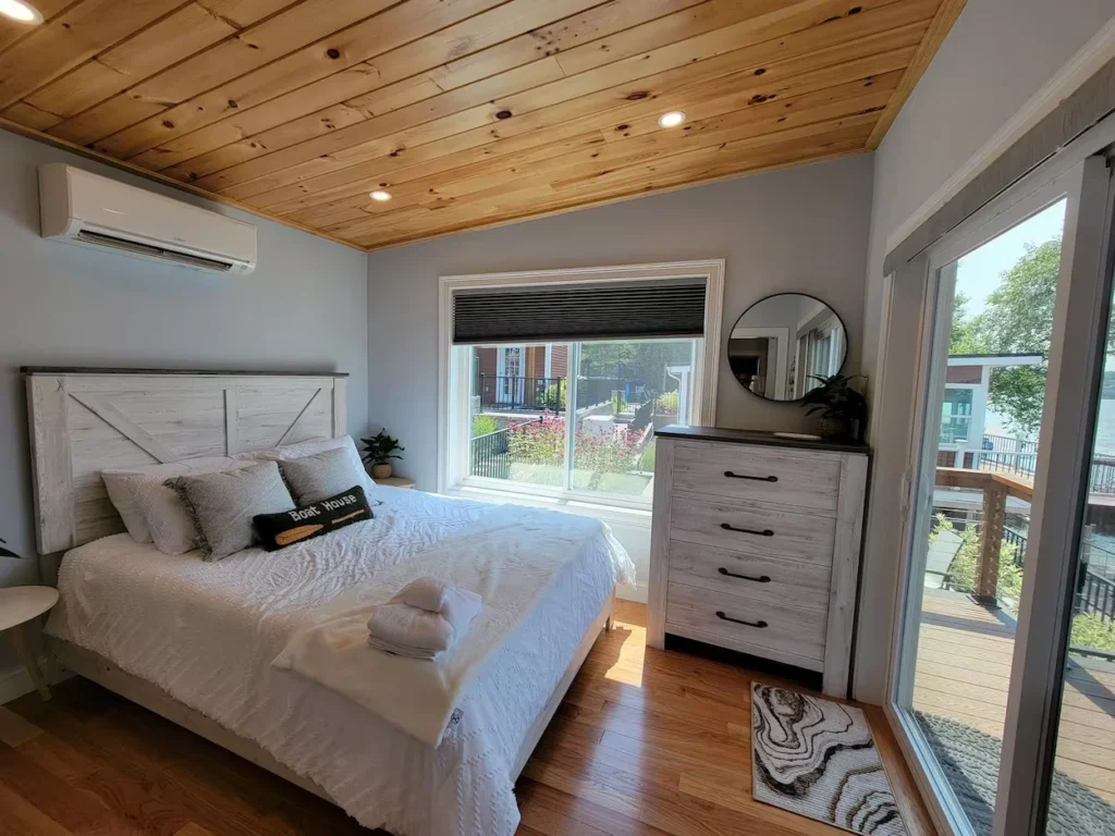 Cabin bedroom with wood-paneled ceiling, white barn-door style bed, and glass door opening to a deck