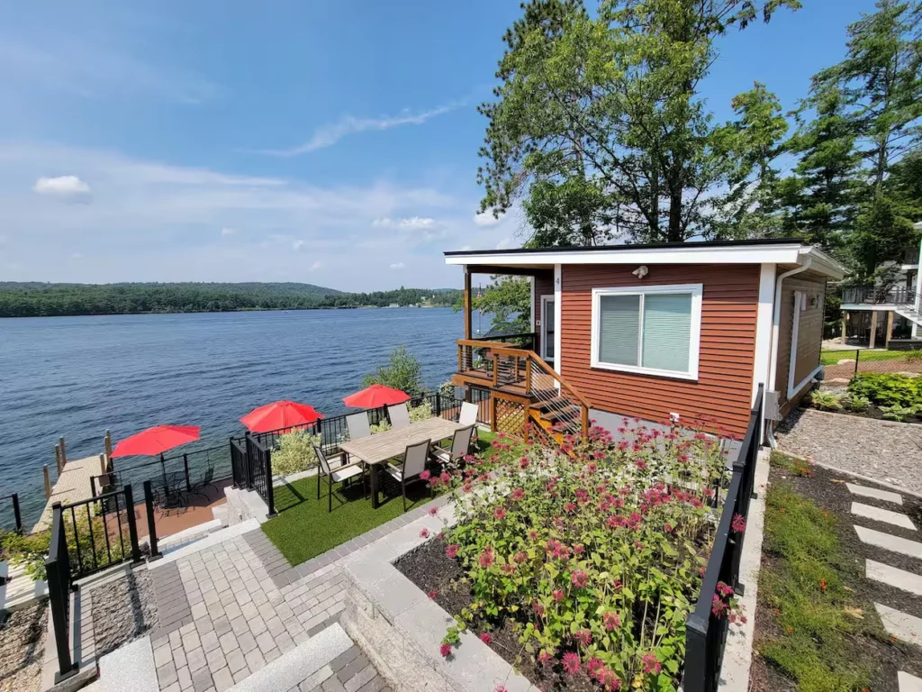 Lakefront brown cabin with an outdoor dining patio, flower beds, and docks under a blue sky