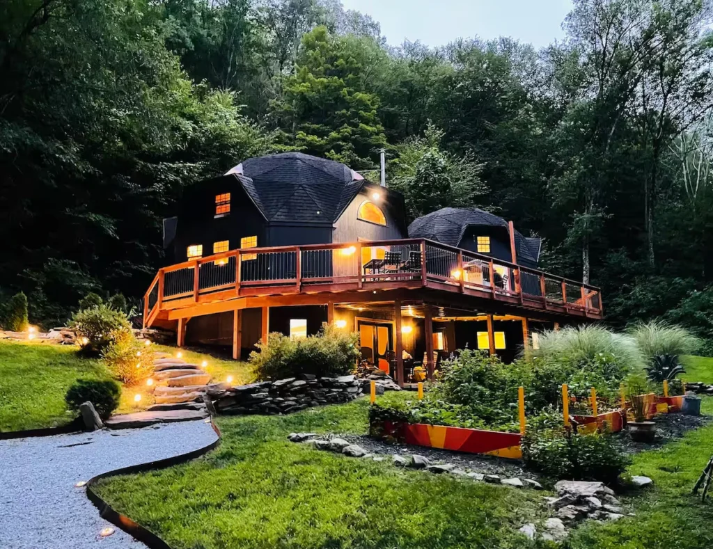 Black geodesic dome house lit up at dusk with a large wrap-around wooden deck and garden