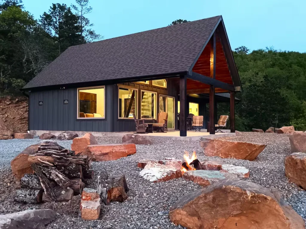 Modern dark chalet exterior with a lit stone fire pit and patio seating at dusk