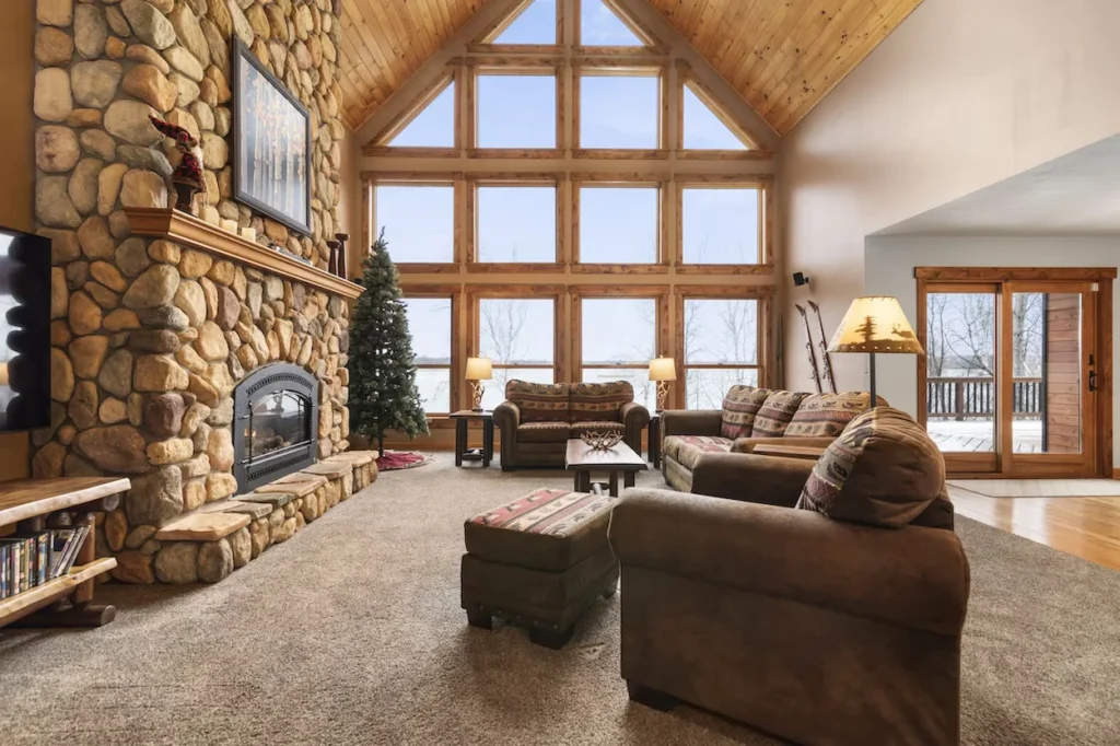 Rustic cabin living room featuring a tall stone fireplace and floor-to-ceiling windows overlooking a lake