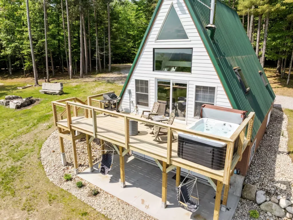 White A-frame cabin with a green roof, elevated wooden deck, and a hot tub