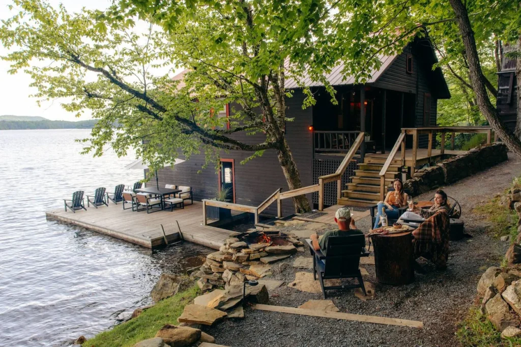Dark wood lakeside cabin in Old Forge, New York, with a spacious deck and people gathered by a stone fire pit