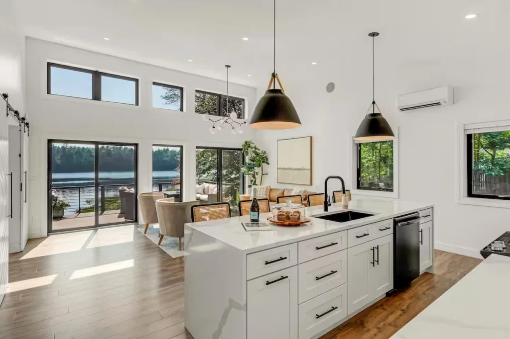 Bright open-concept kitchen with a white island, black pendant lights, and large windows facing a lake