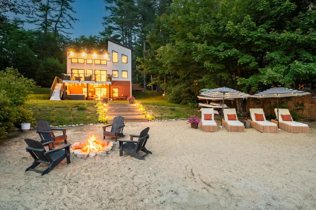 Modern lakeside home at dusk with a sandy beach, campfire ring, and lounge chairs