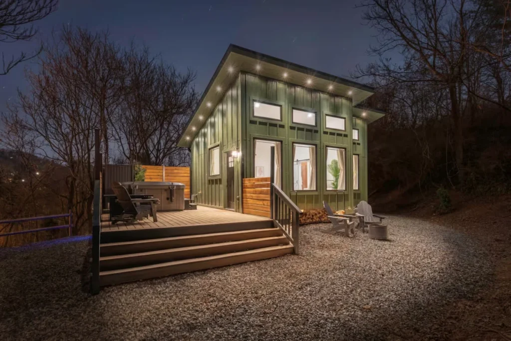 Green modern cabin lit up at night with a hot tub on the wooden deck