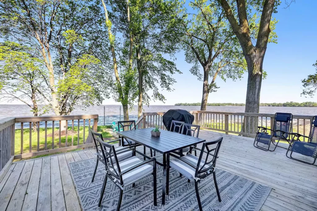 Wooden deck with an outdoor dining table, chairs, and lounge seating overlooking a wide lake