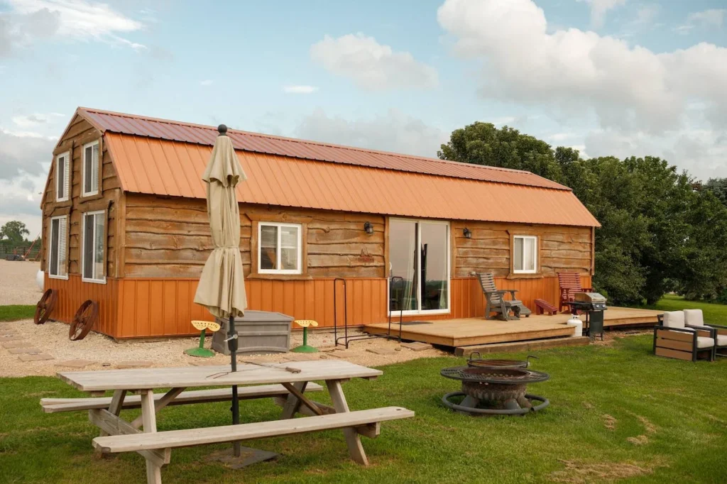 Log cabin with an orange gambrel roof, wooden deck, picnic table, and fire pit on a grassy lawn