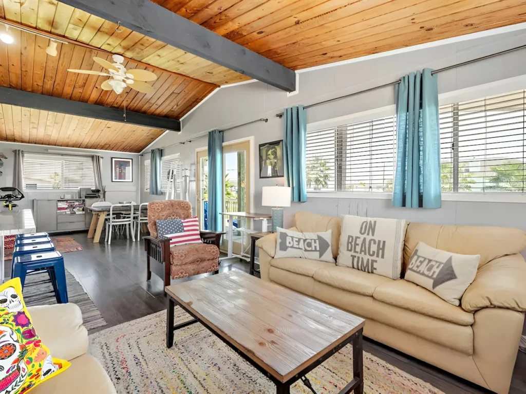 Beachy living room with a tan sofa, wood-plank ceiling, and large windows with blue curtains