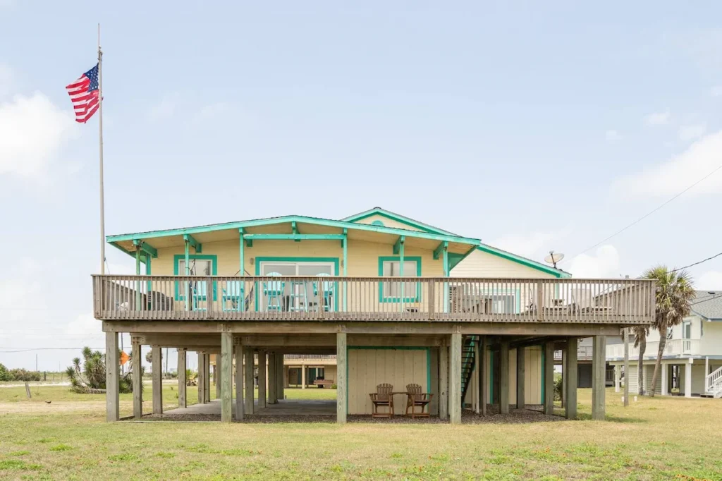 Yellow and teal beach house on stilts with a wide wooden deck and an American flag