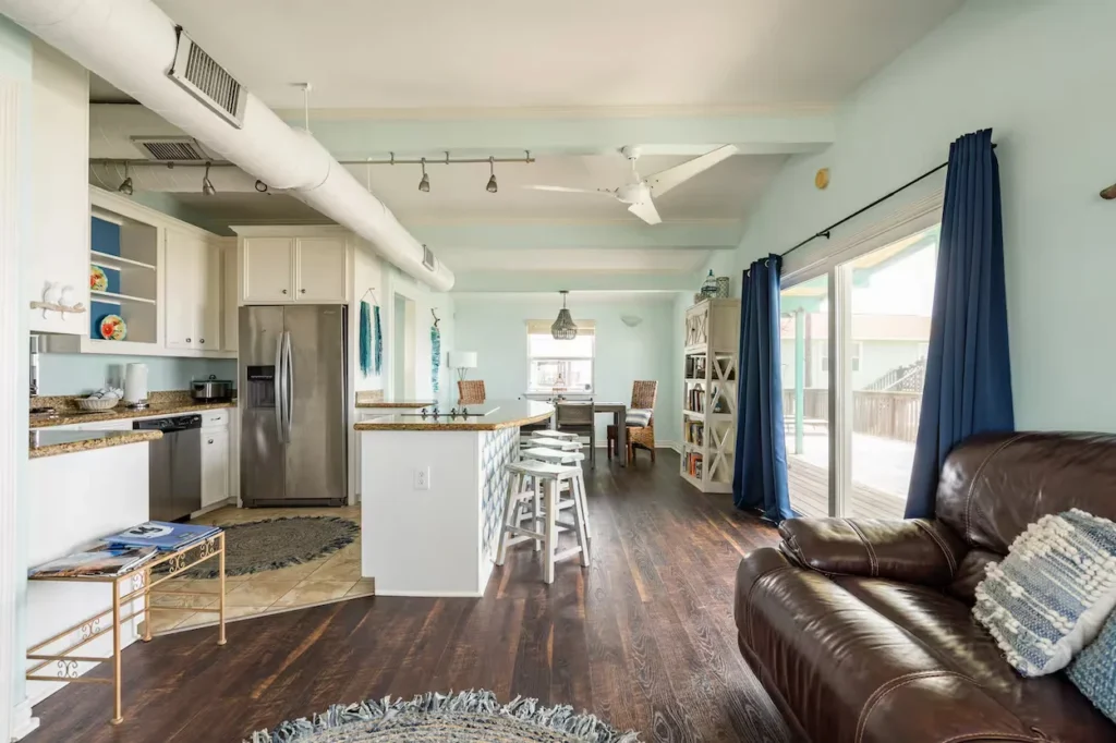 Open-concept beach house interior with light blue walls, dark hardwood floors, white kitchen cabinets, and a brown leather sofa
