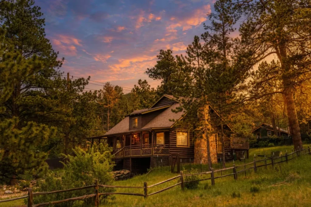 Dark log cabin with a stone chimney and wooden fence on a grassy hill at sunset