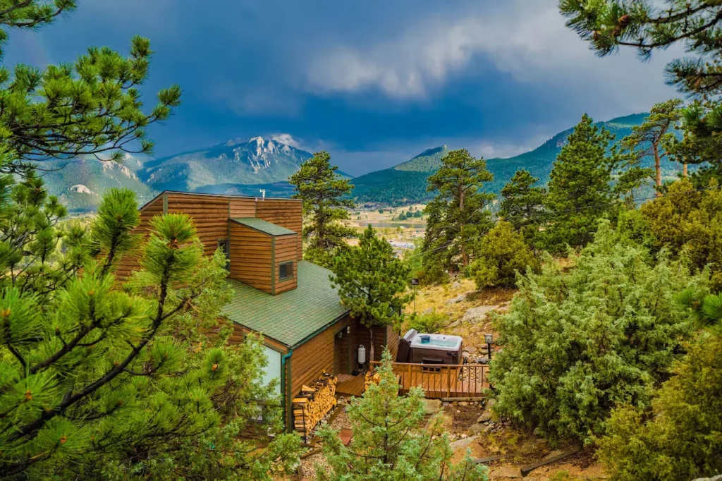 High-angle view of a wooden cabin with an outdoor hot tub facing distant mountains under dark clouds