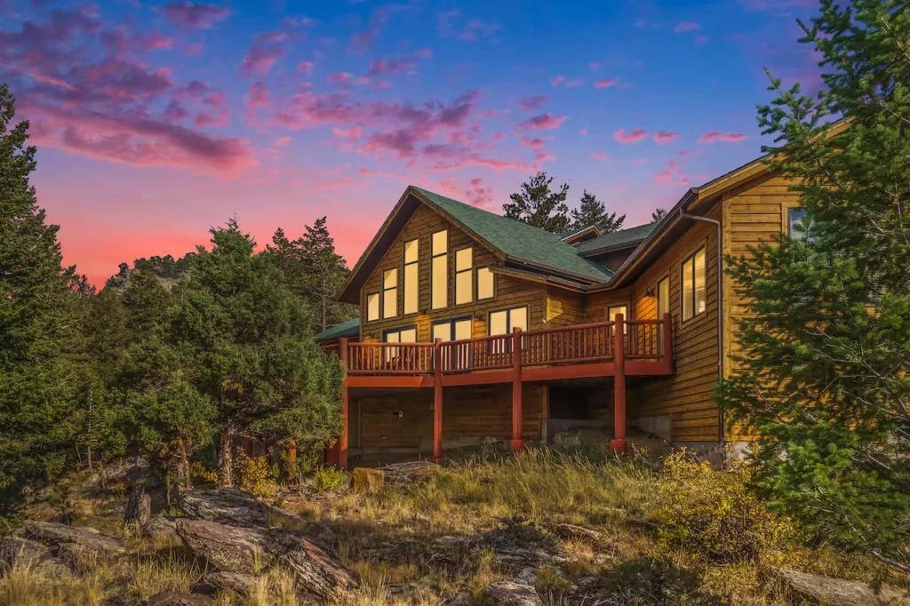 Large wooden mountain cabin with a raised deck at sunset surrounded by pine trees