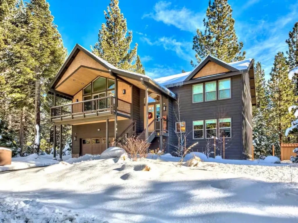 Large two-story dark wood mountain cabin with a covered deck surrounded by deep snow and pine trees