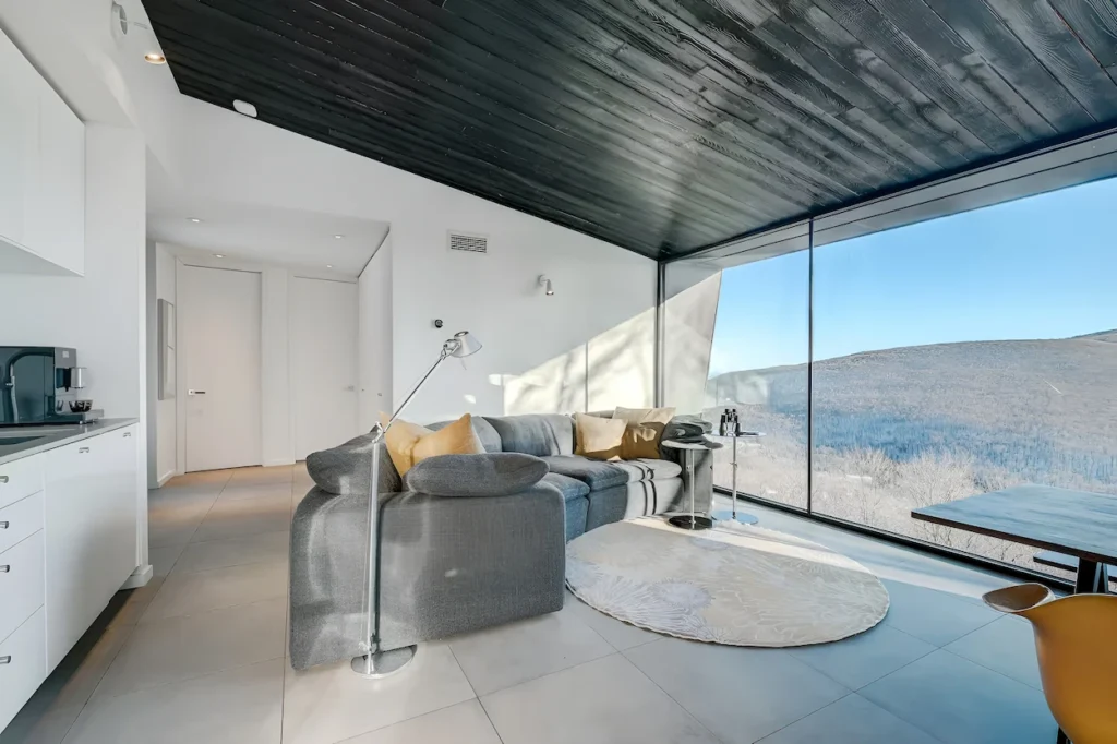 Minimalist cabin living room with a grey sectional couch, black wood ceiling, and panoramic mountain views
