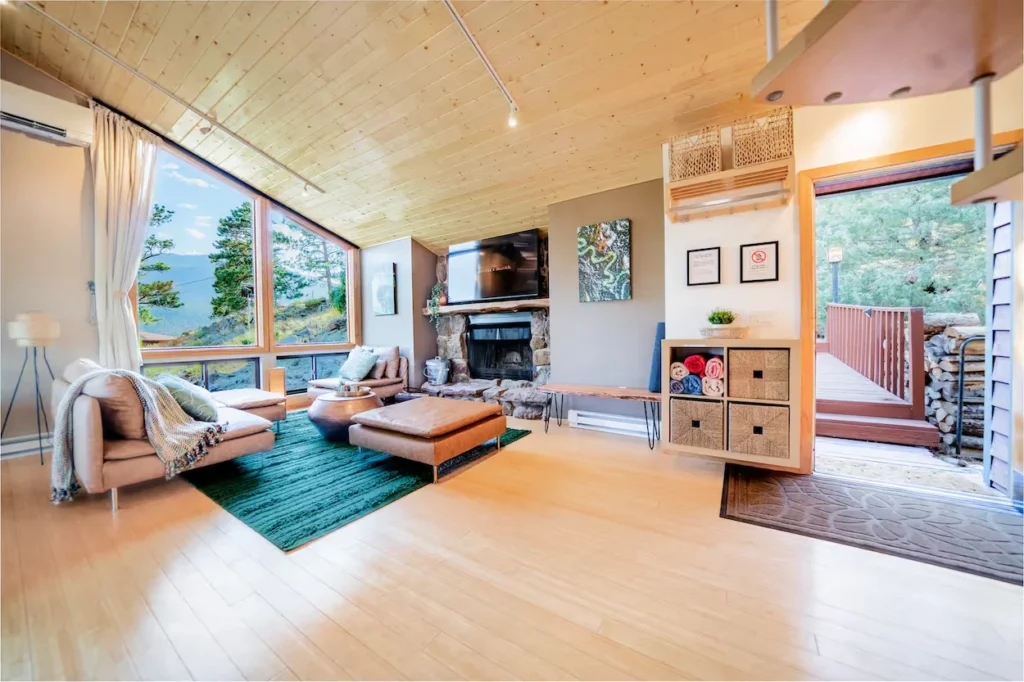 Bright cabin living room with a stone fireplace, light wood ceiling, and large picture windows