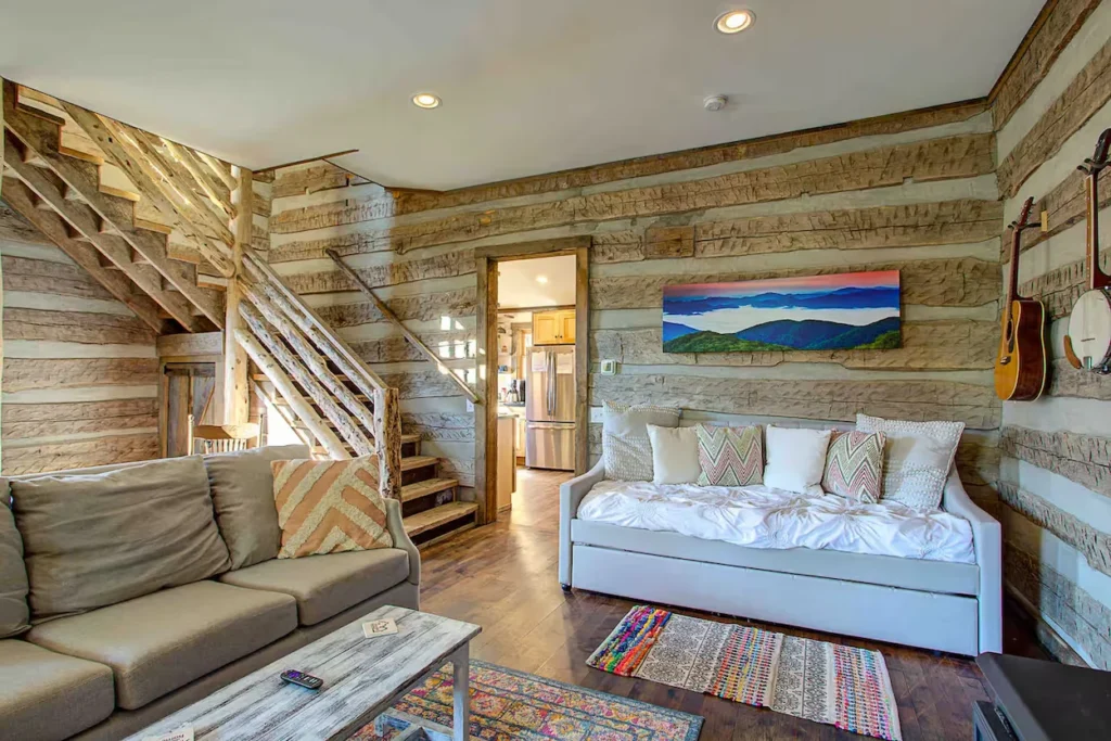 Log cabin living room with log-style stairs, chinked log walls, a grey sofa, and a daybed under a landscape painting