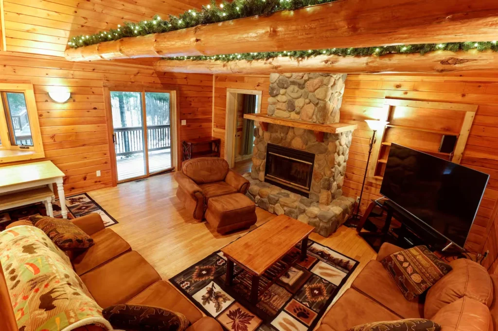 Log cabin living room with a stone fireplace, brown sofas, and string lights on overhead logs
